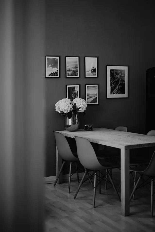 Black and white photo of a dining table with four chairs, a vase of flowers, a camera, and framed landscape pictures on the wall.