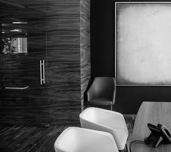 Black and white photo of a modern office corner with a glass wooden door, two white chairs, a gray armchair, a table with a phone, and a blank framed artwork on the wall.