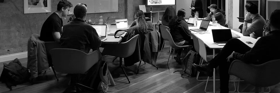 Black and white photo of a group of people seated around tables working on laptops in a modern office setting.